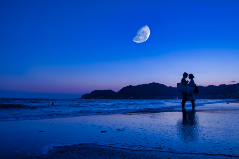 A couple hugging on the beach during dusk with the moon partially visible in the blue sky.