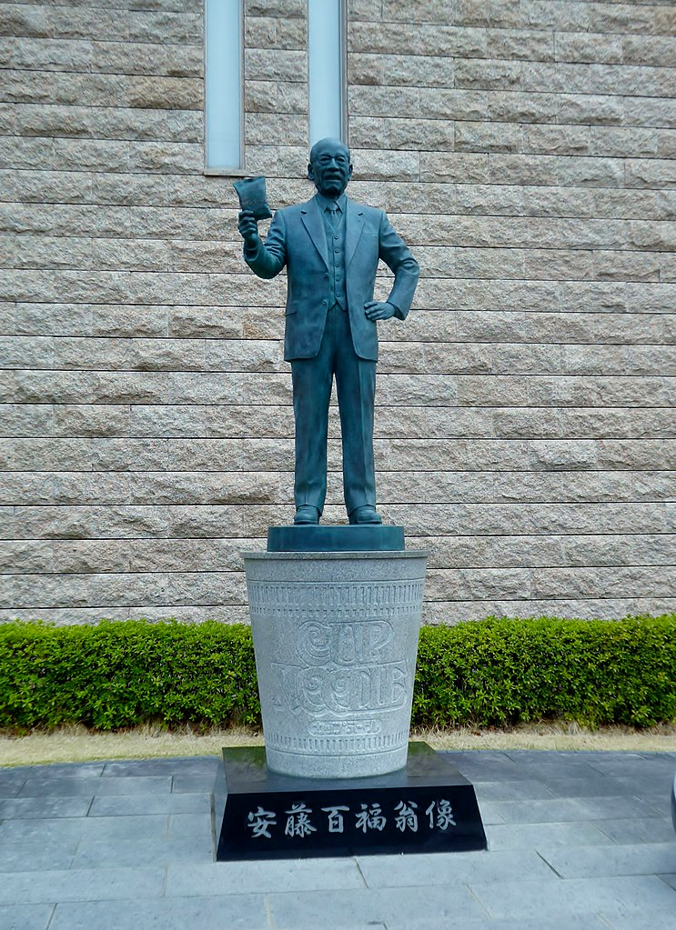 A statue of Momofuku Ando holding up a packet of Nissin Chicken Ramen standing on a giant cup of Nissin Cup Noodles.