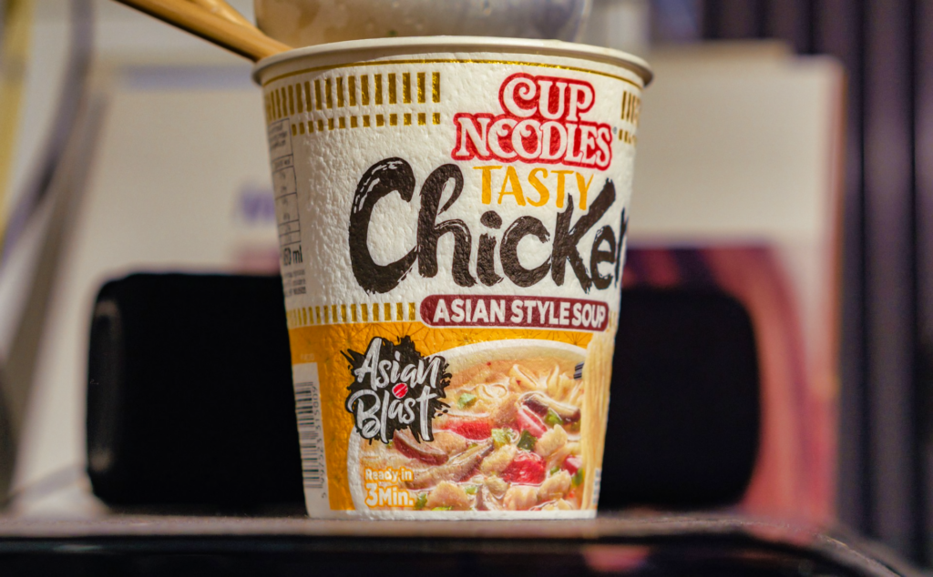 A cup Asian style soup flavored of Nissin Cup noodles with the lid open and pair of chopsticks sticking inside.