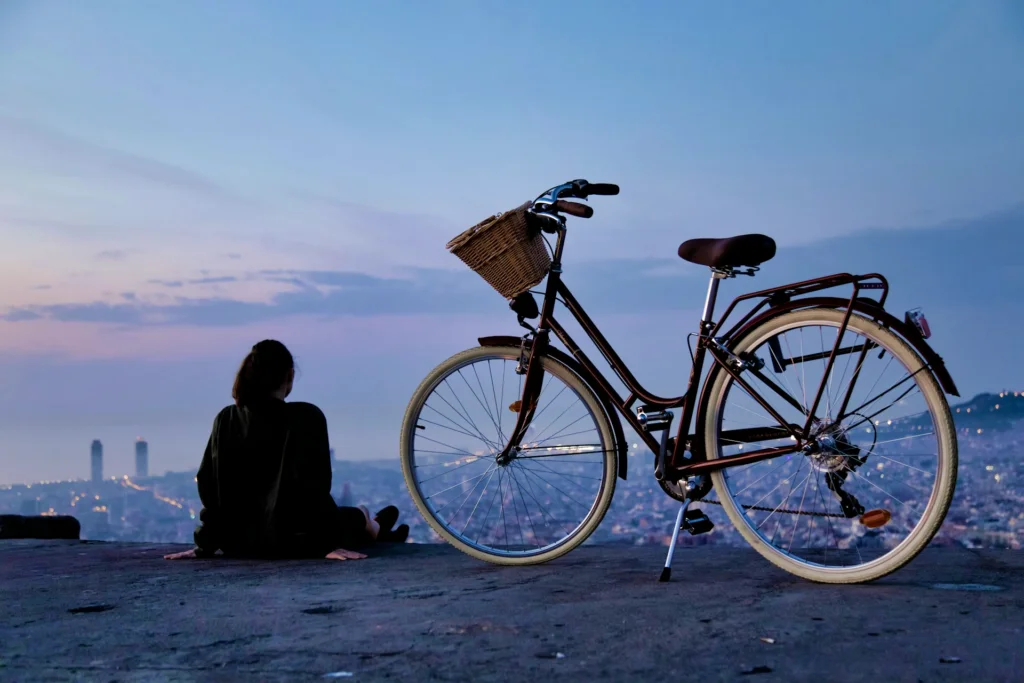 A Spanish girl sitting on a hill overlooking Barcelona with a mamachari style bicycle next to her.