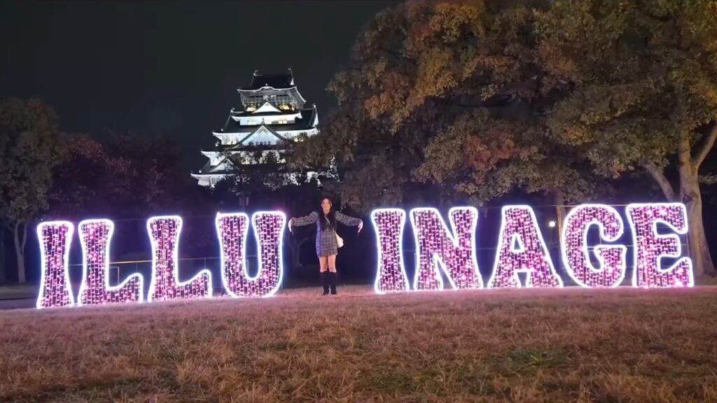 Hazunats in front of the Illuminage sign attempting to make an "M" character with her arms.