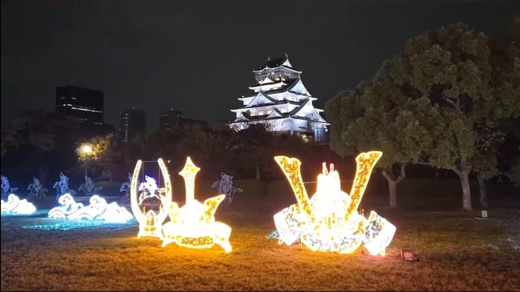 A view of Osaka castle at night from behind 2 samurai helmet illuminations.